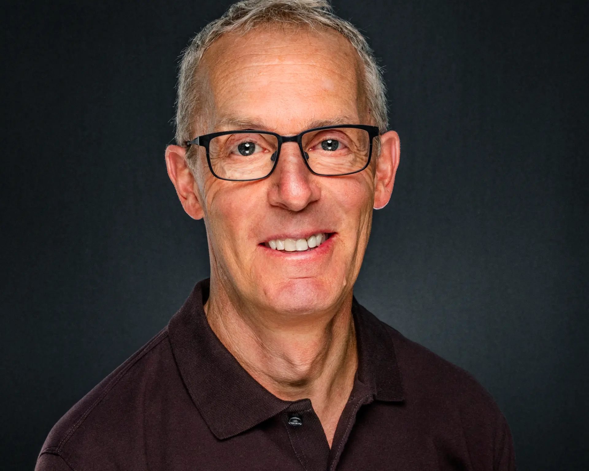 Studio headshot of a smiling man in glasses wearing a dark polo shirt