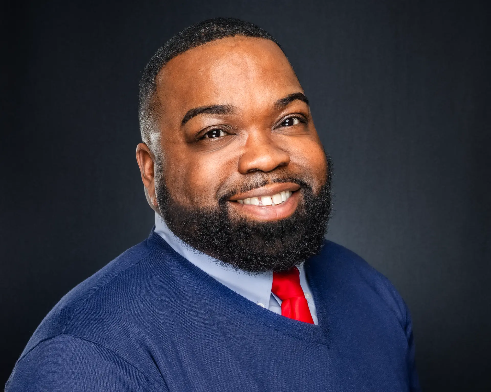 Studio headshot of a smiling professional man in a blue sweater and red tie