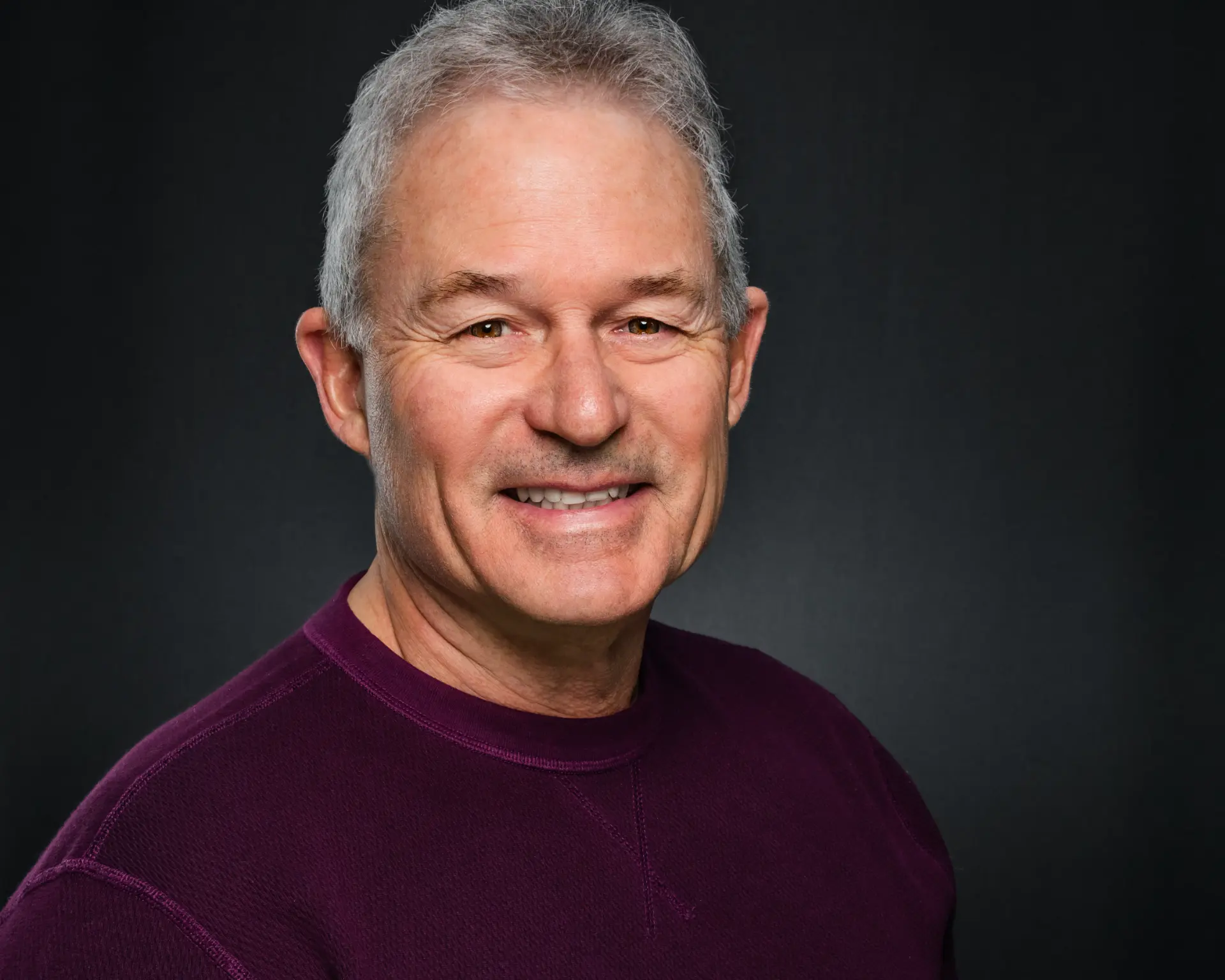 Studio headshot of a smiling man with silver hair in a plum sweater